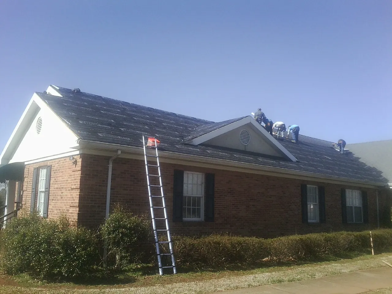Crew tearing shingles off a brick ranch roof with a ladder set — Emergency Roof Repair work by our crew in Bay City